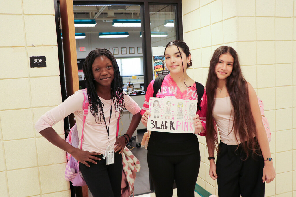 Three girls standing together in a hallway, holding a sign that says "Black Pink" with images of women.