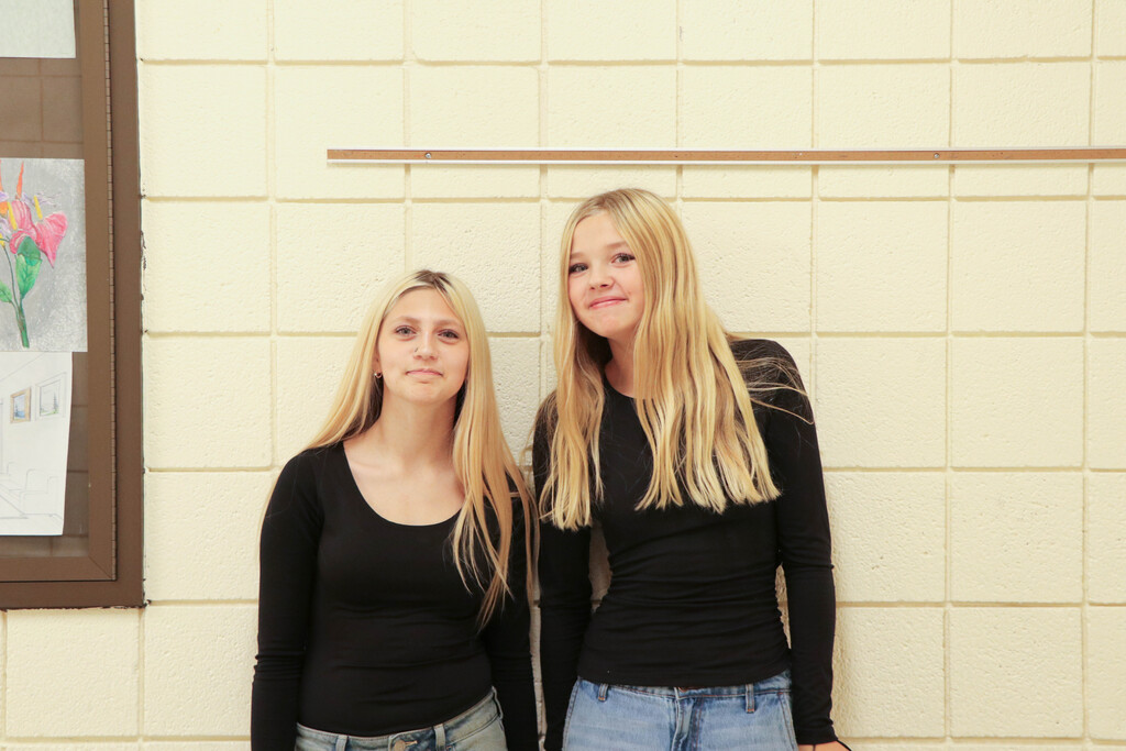 Two women stand together in front of a light-colored wall; one has blonde hair, the other has dark hair.
