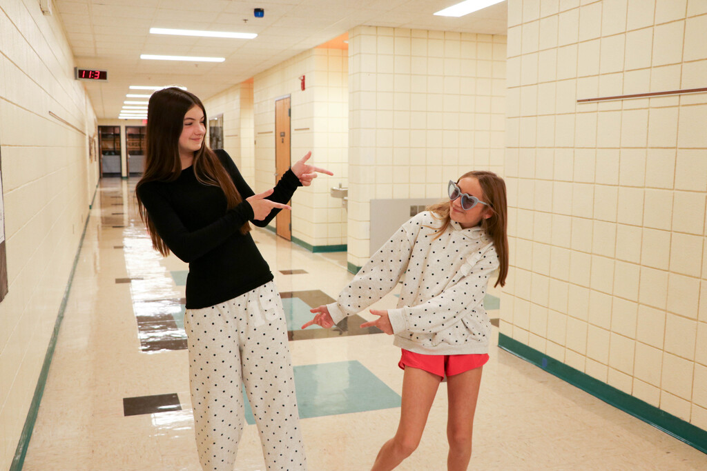 Two girls stand in a school hallway with tiled walls and white ceiling lights, one wearing sunglasses.