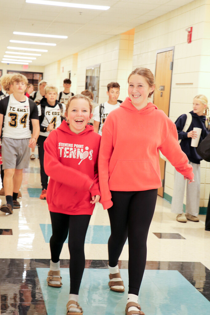 Two girls in hoodies, one red, one orange, walking in a hallway with other students.
