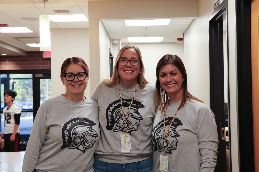Three women wearing gray sweatshirts with helmet designs stand together in a school hallway. One wears glasses, another a lanyard.