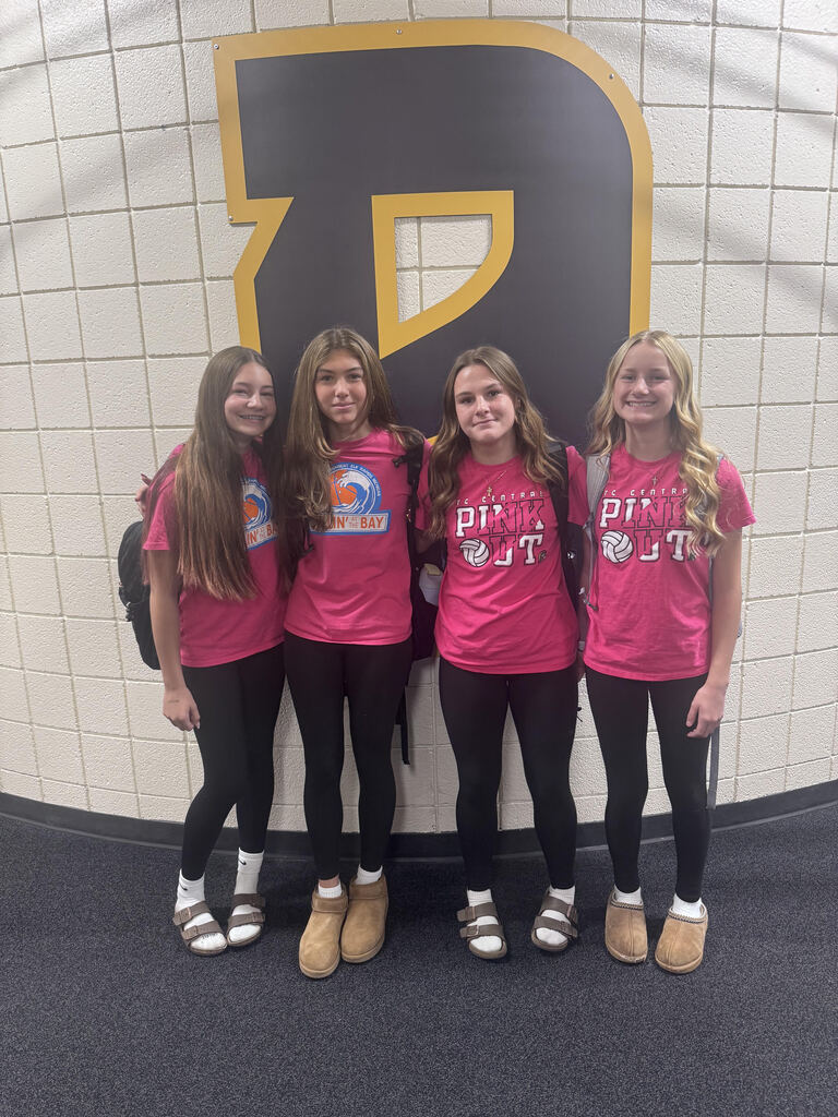 Four young girls wearing matching pink shirts and black leggings pose together, standing in front of a wall.