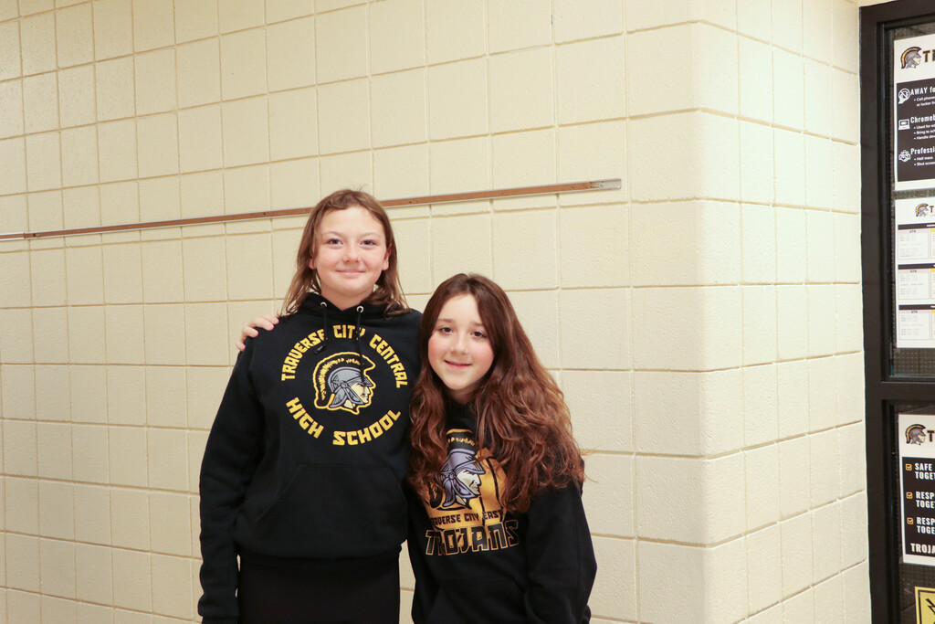 Two girls in black sweatshirts stand next to each other against a white wall with bulletin boards.