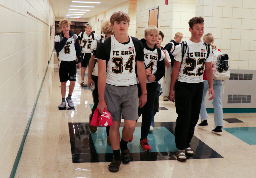 Students wearing numbered shirts walk in a school hallway with white walls, blue floor, and ceiling lights.