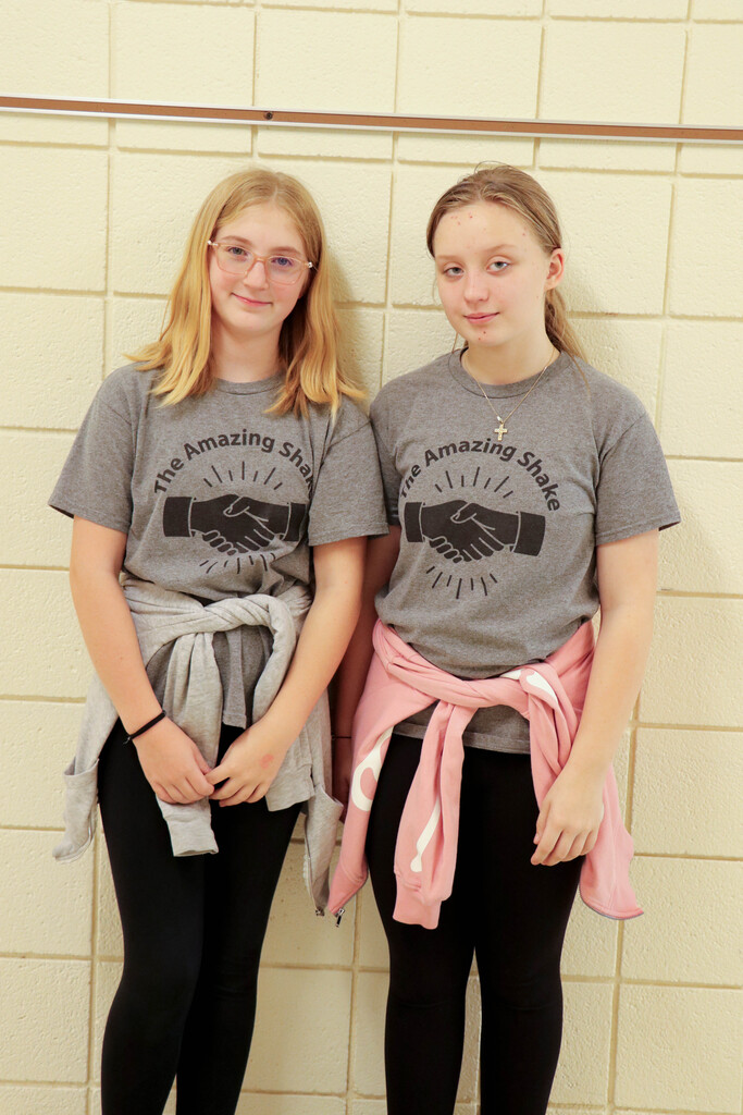 Two girls stand close to each other wearing gray shirts and black leggings. One girl wears glasses and has blonde hair.