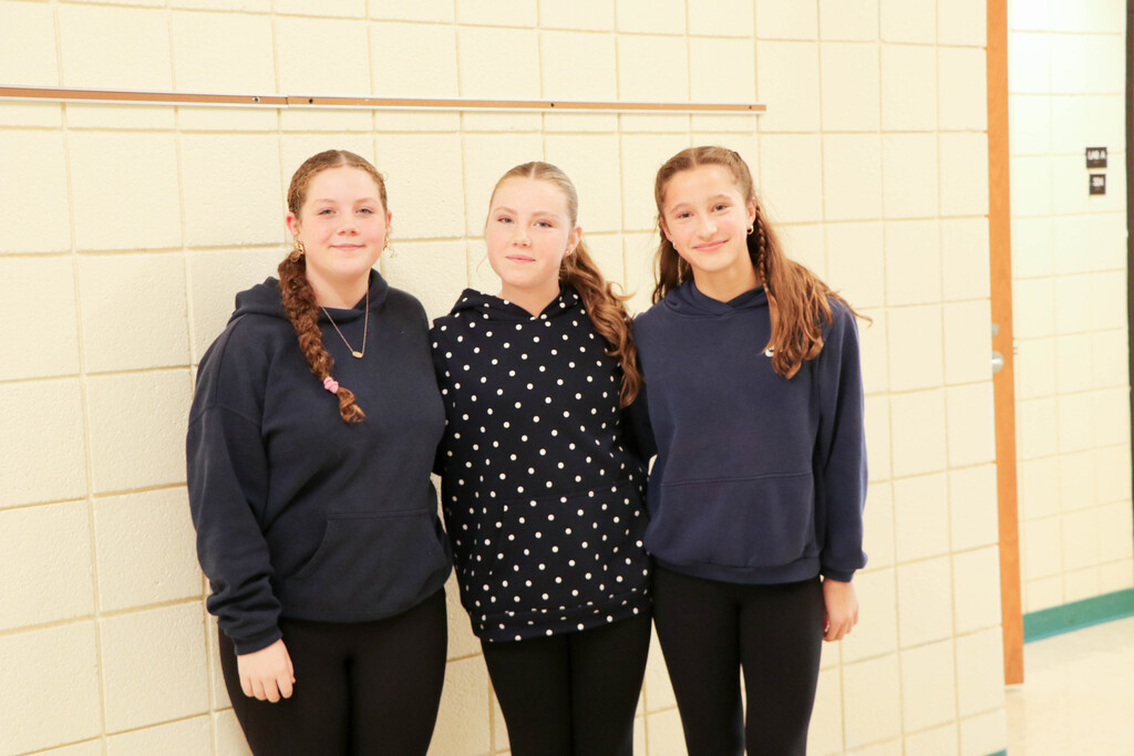 Three girls standing next to each other. One wears a black hoodie, another a polka-dot shirt, and the last a navy blue sweatshirt. They are in front of a wall.