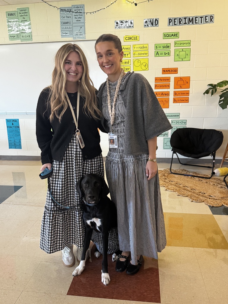 Two women stand with a black and white dog. One holds a leash. Behind them, a white board and green plant are visible.