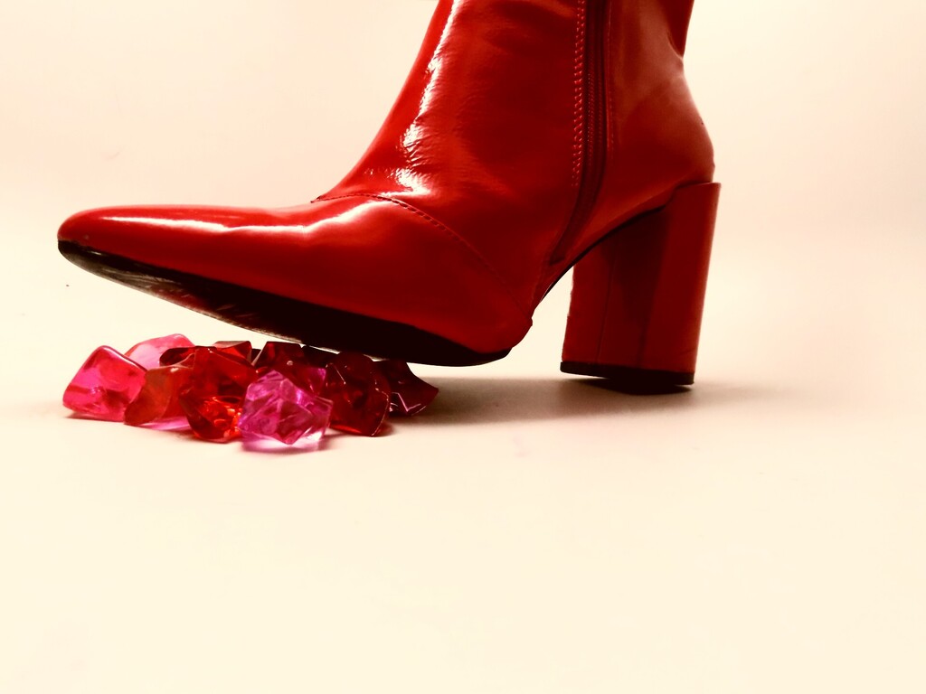 Red high-heeled boot stepping on scattered red and pink crystals against a white background.
