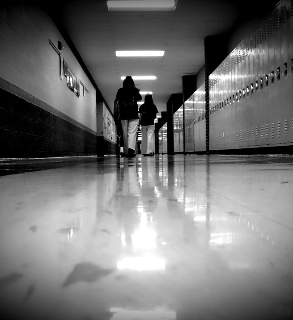Two people in front of lockers walk down a hallway with white tile floor and white lights on ceiling.
