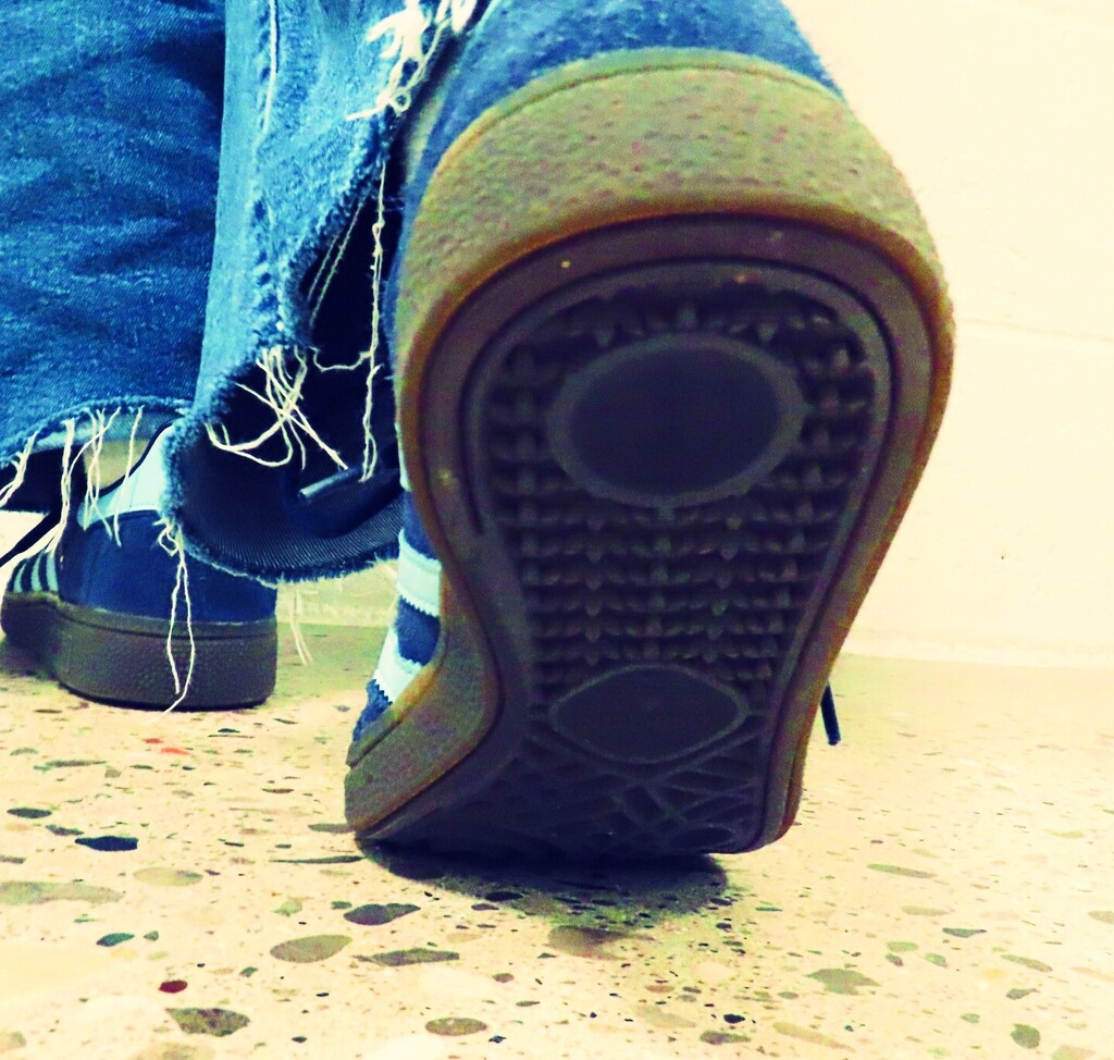A pair of worn blue jeans and a shoe with a worn-out sole. The shoe is positioned on a speckled floor.