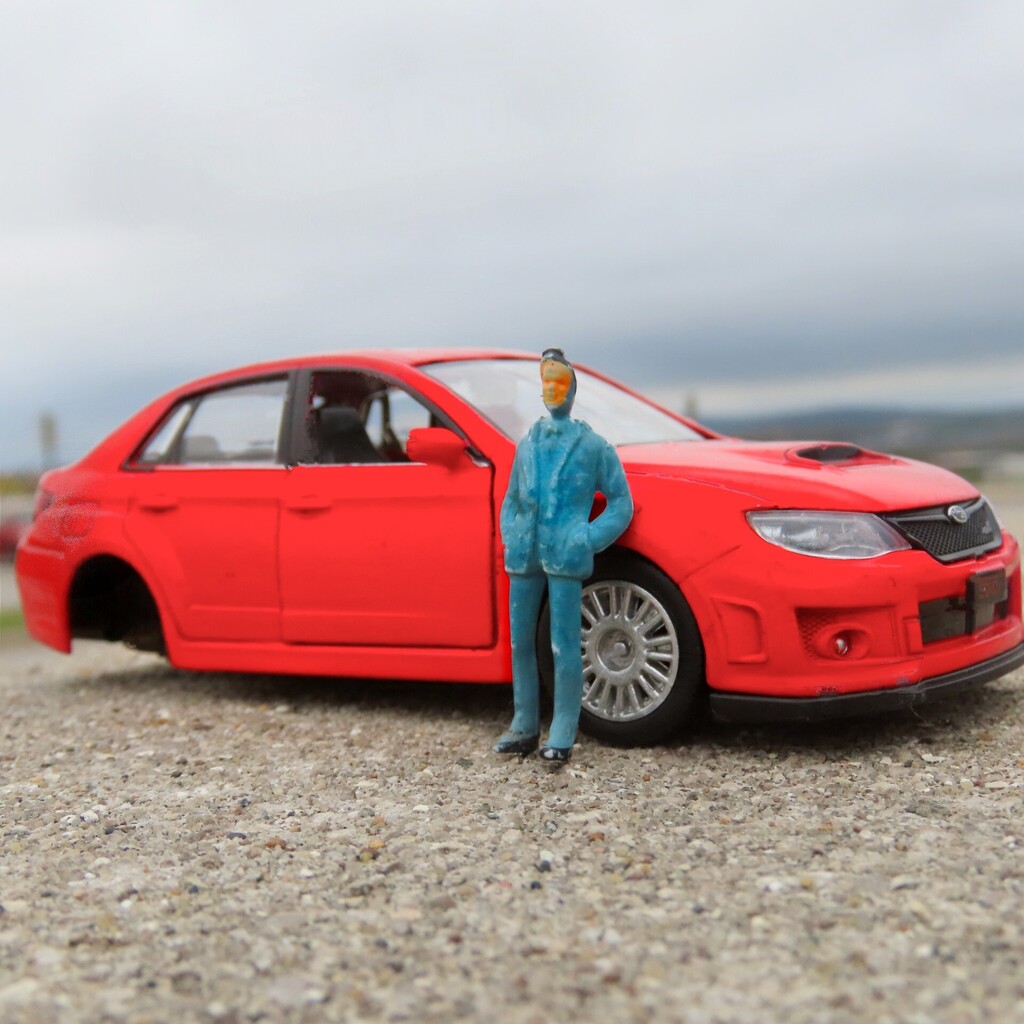 Miniature model of a red car with a figure of a man in blue suit standing next to it on gravel.