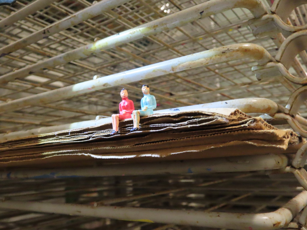 Two small figurines sit on a stack of cardboard; one wears a red outfit and the other a blue one.
