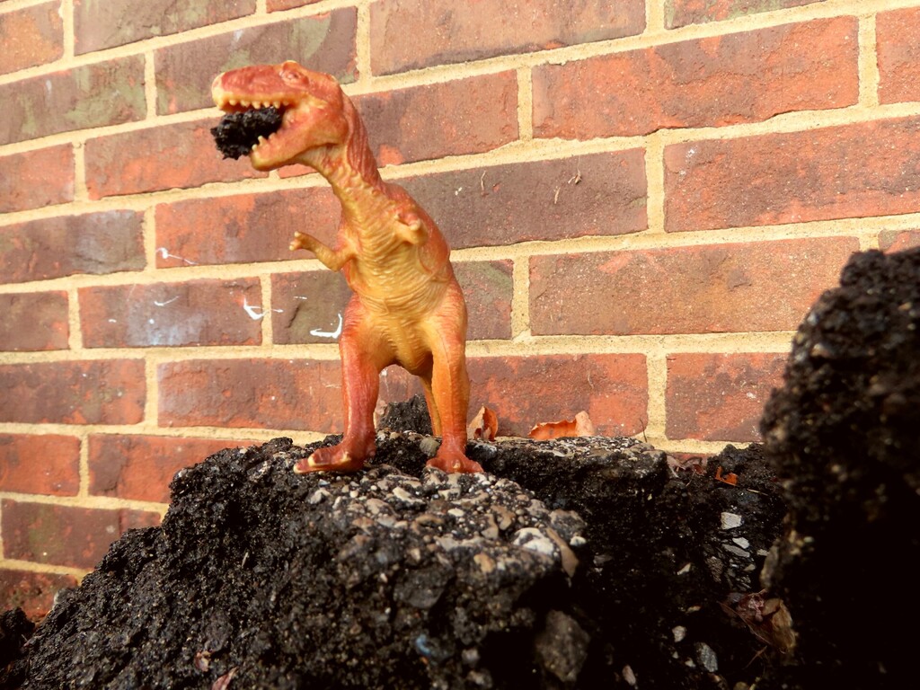 A toy dinosaur stands on a pile of rocks in front of a red brick wall.