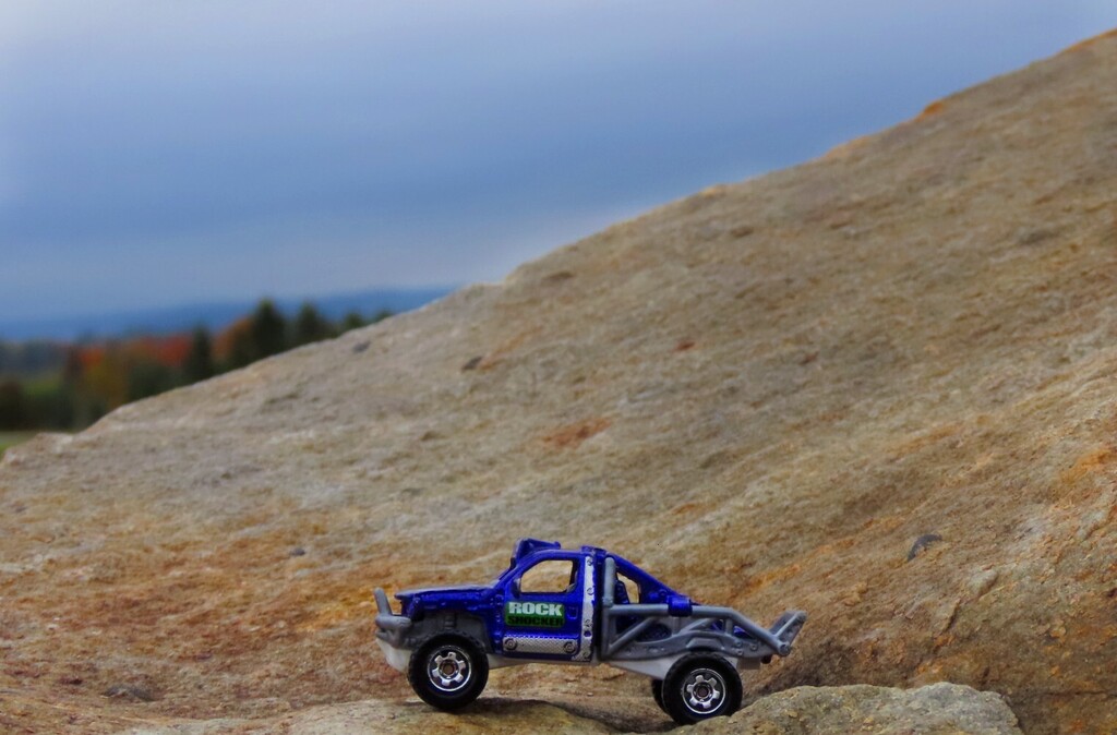 A blue toy truck with a large tire is parked on a steep rocky hillside. Trees and mountains are in the background.