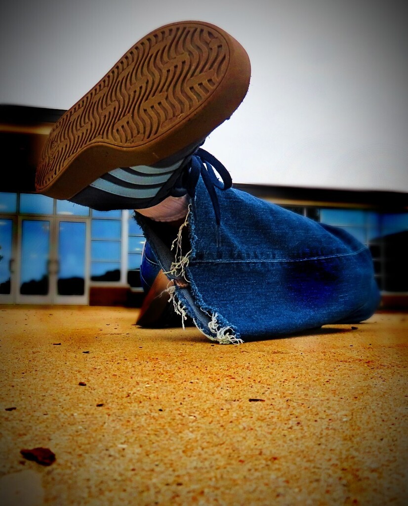 Person in jeans with a torn knee and a sneaker showing, lying on a concrete floor with windows behind.