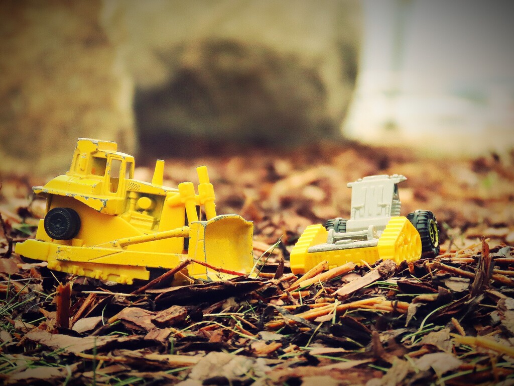 Two yellow toy bulldozers on fallen leaves, one facing left, one facing right, on a concrete background.