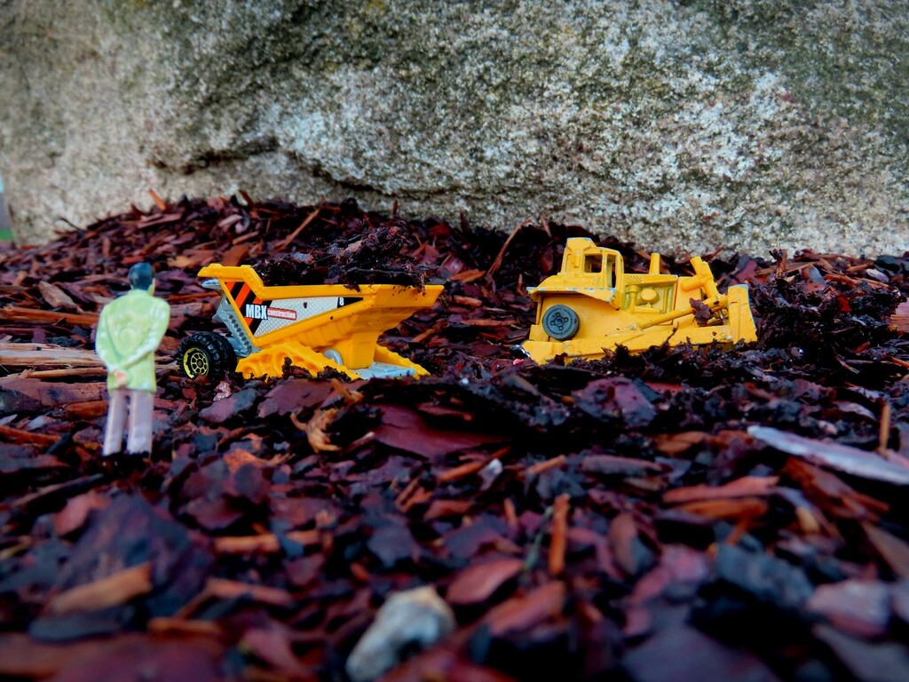Two yellow toy construction vehicles and a tiny figure standing next to them on a bed of brown mulch.