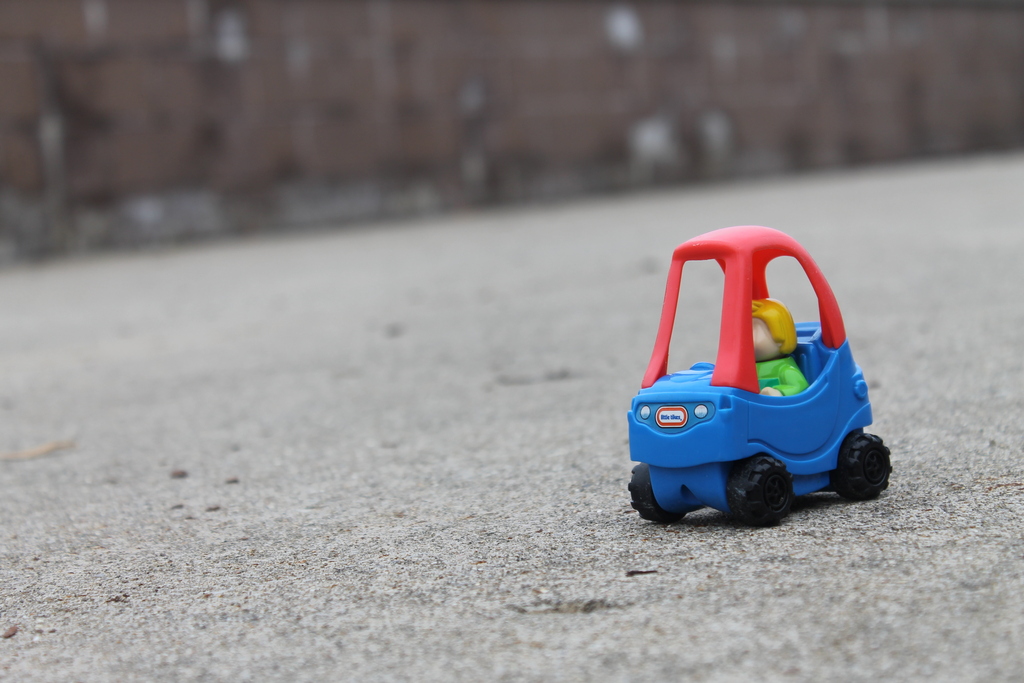 Small blue toy car with a person figure sits on a road in front of a fence.