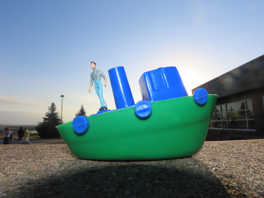 A green toy boat with blue cylindrical pieces on a concrete surface, with a small figurine standing on top, near a building.