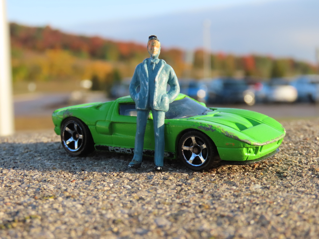 A green toy sports car and a small figurine of a person in a blue coat. The background shows a parking lot with cars parked.
