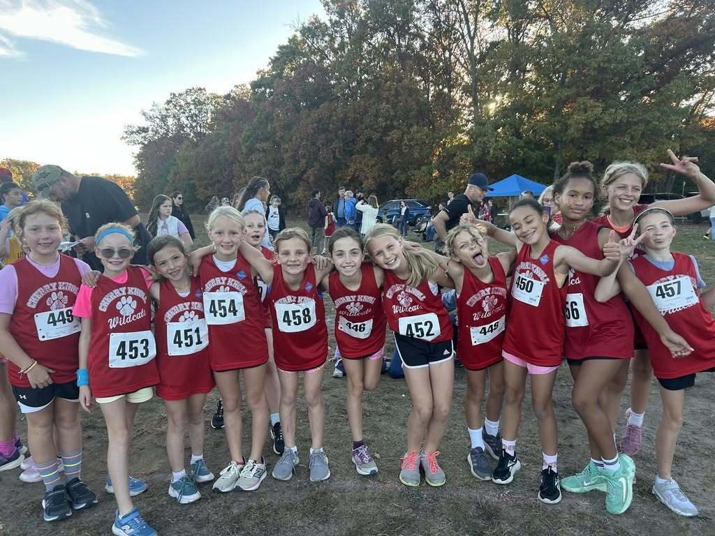 Several children wearing red and white athletic uniforms with numbers, standing in a row in a field.
