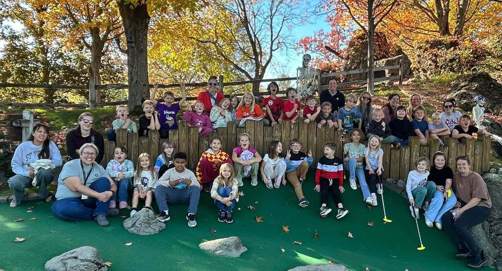 A group of students and adults are seated and standing on a green surface in an outdoor area.