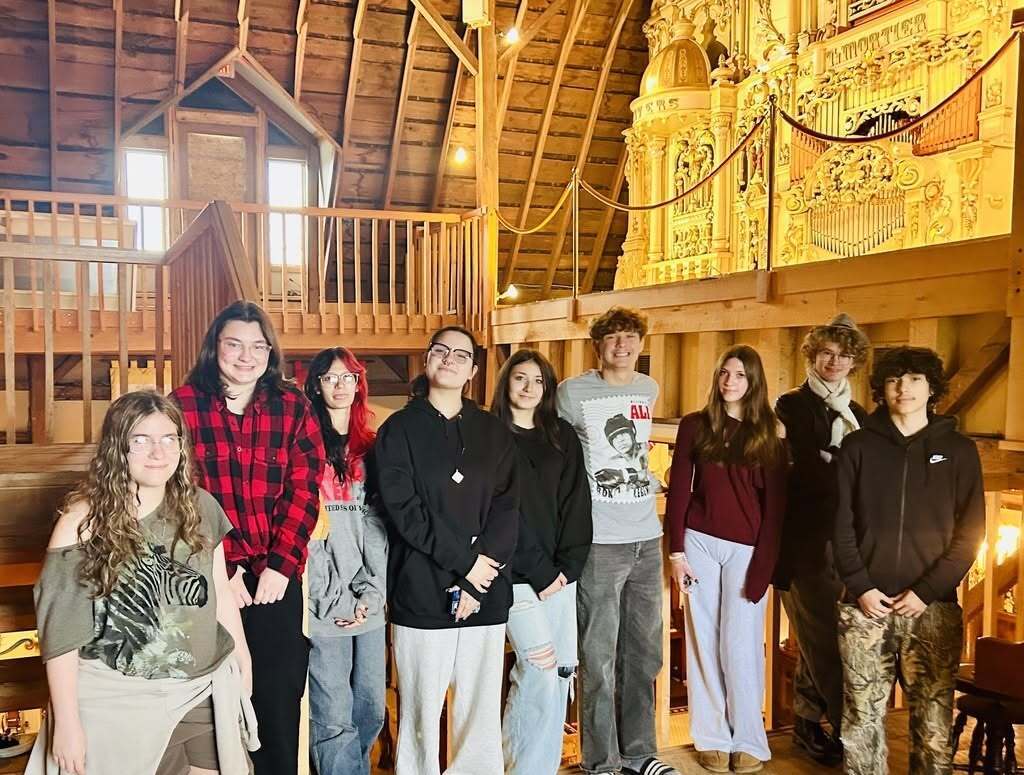 A group of students stands inside a wooden building with windows and a balcony.
