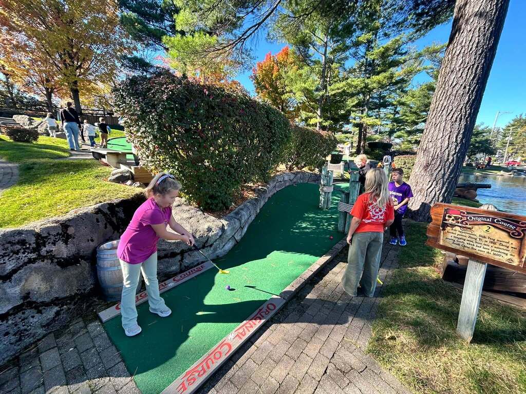 People play mini golf on a course with green putting surfaces. Trees and bushes line the edges.