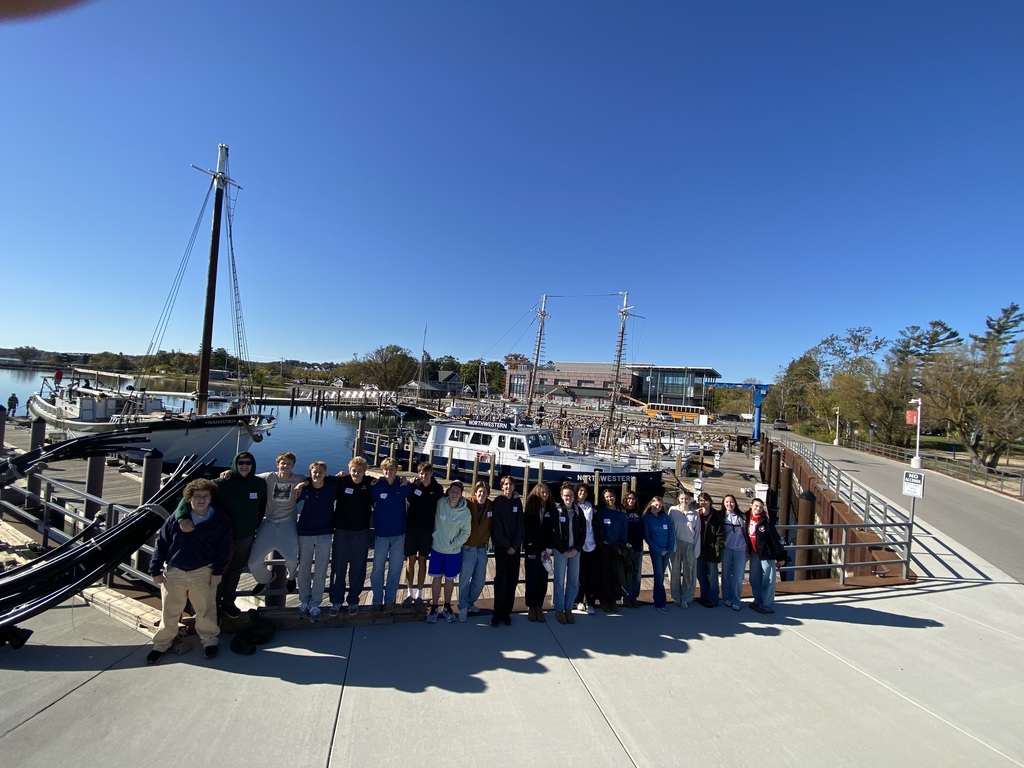 A group of students and teachers pose on a dock near a marina with sailboats.