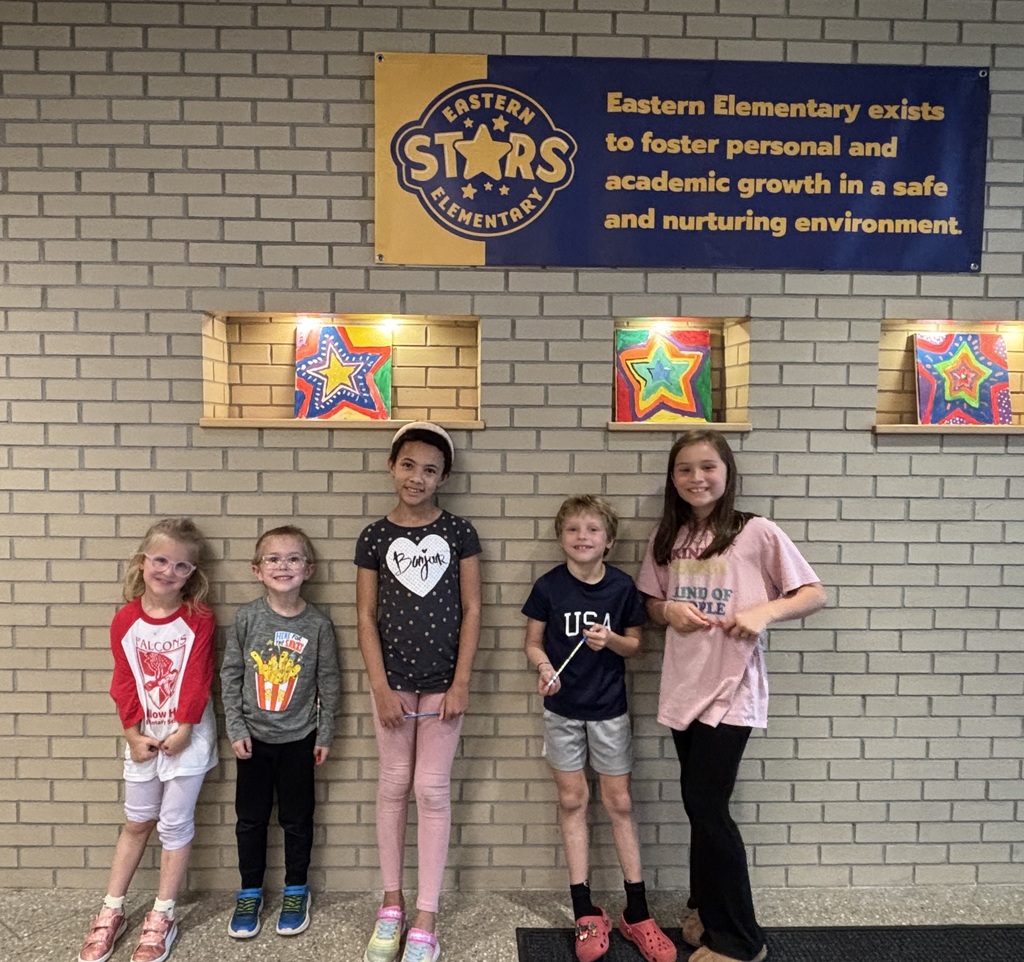 Five children stand next to each other in front of a brick wall with artwork and a banner.