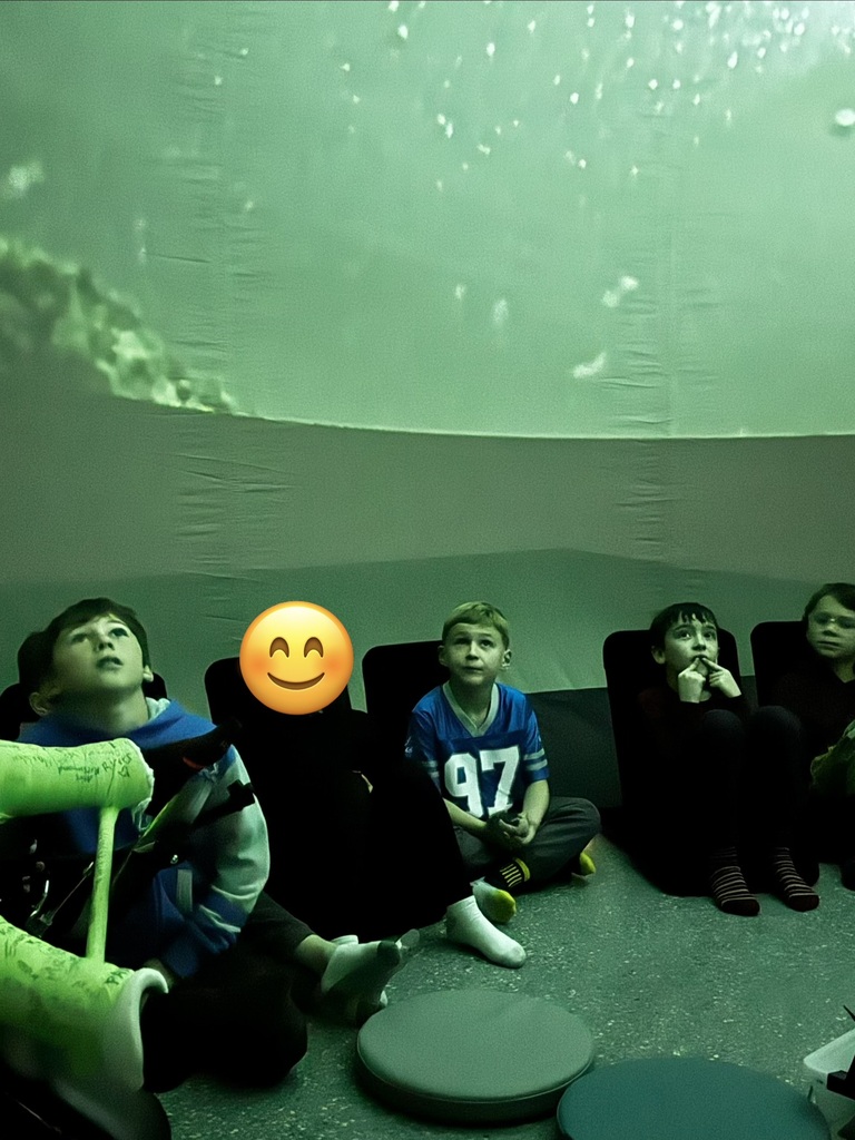 Several children sit on floor, watching an underwater scene projected on wall. One child wears a smiley face mask.