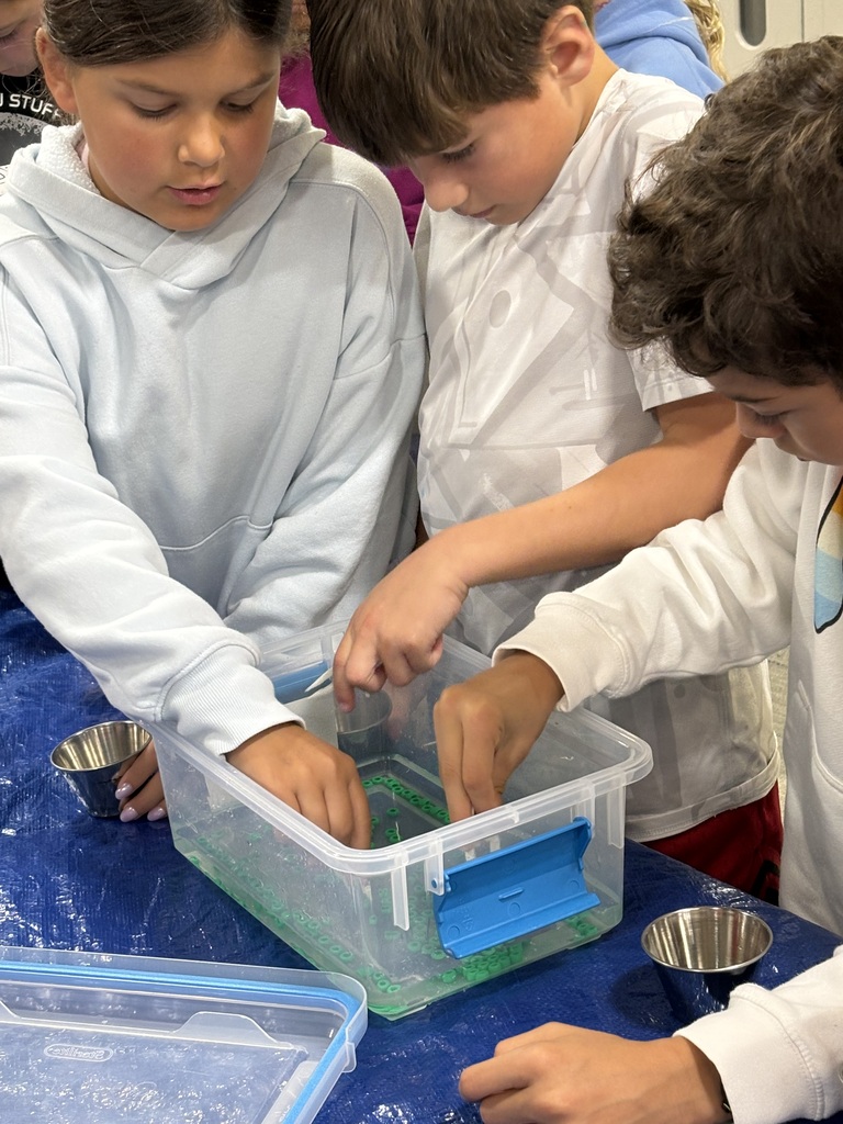 Three students use clear containers to observe liquid and solid objects. One container has a blue liquid and green object.