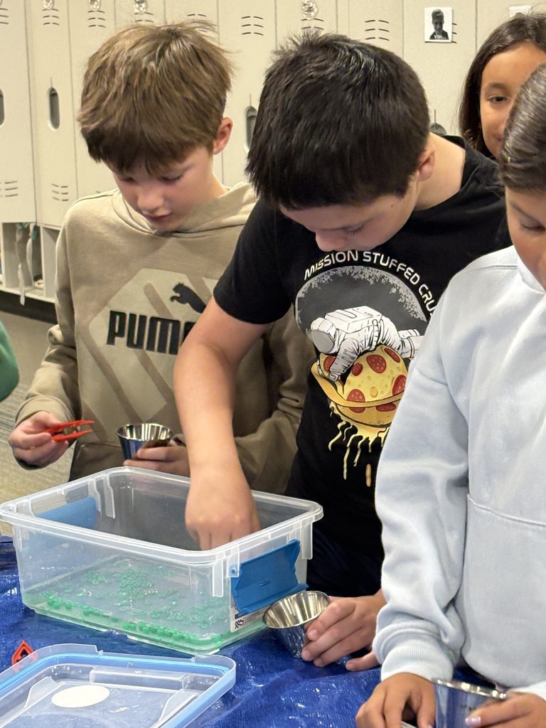 Three students work on a science experiment with containers, tweezers, and a blue table.