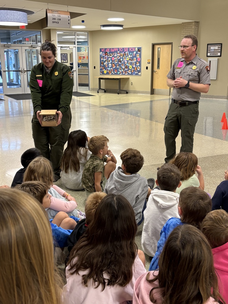 Two adults in uniform holding boxes with children sitting on the floor in front of them.