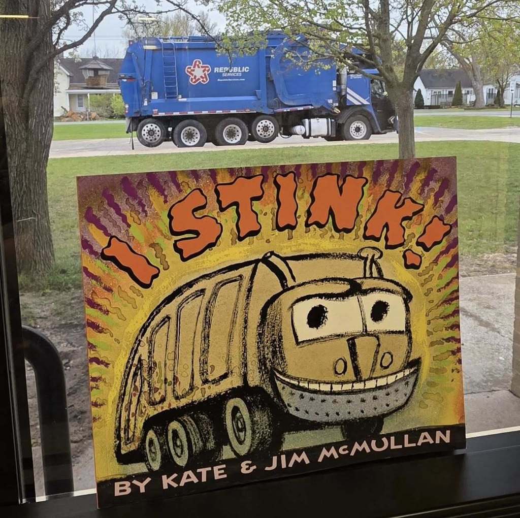 The book "I Stink" in a window sill with a garbage truck behind it.