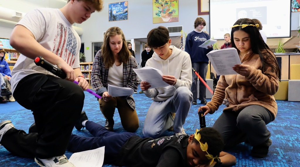 Students kneeling around a downed Caesar in a theater workshop