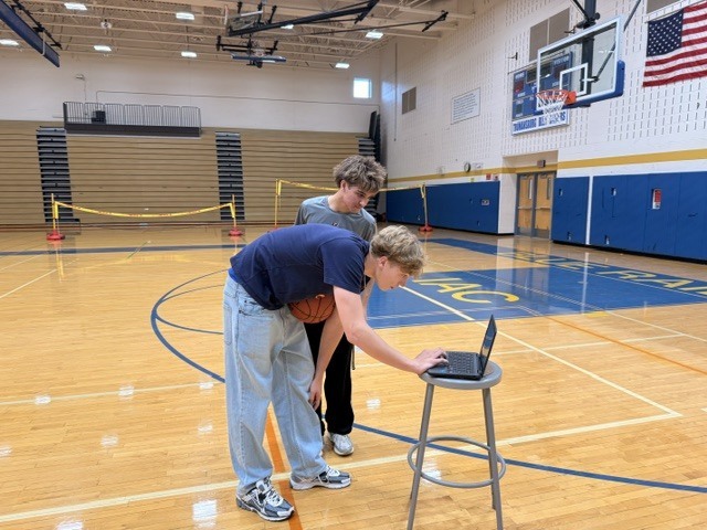 Students in a gymnasium leaning over to look at a laptop