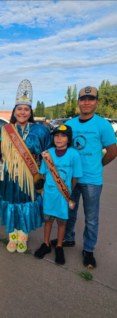Three people stand in a row; a woman and a child wear sashes, and a man stands beside them. The woman wears a blue dress and a white hat. They stand in a parking lot.