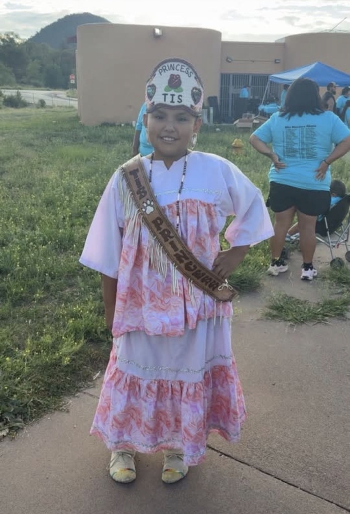 Child stands on path, wearing a sash and traditional dress. A building and grassy area in the background.