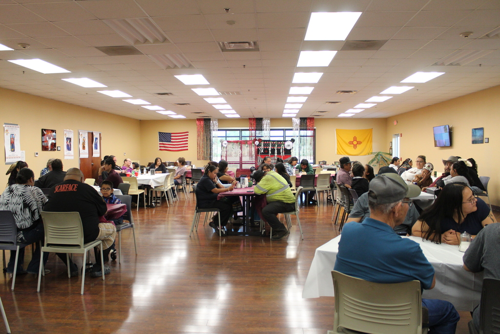 People sit at tables in a room with yellow walls and white ceiling lights. A flag hangs on the wall.