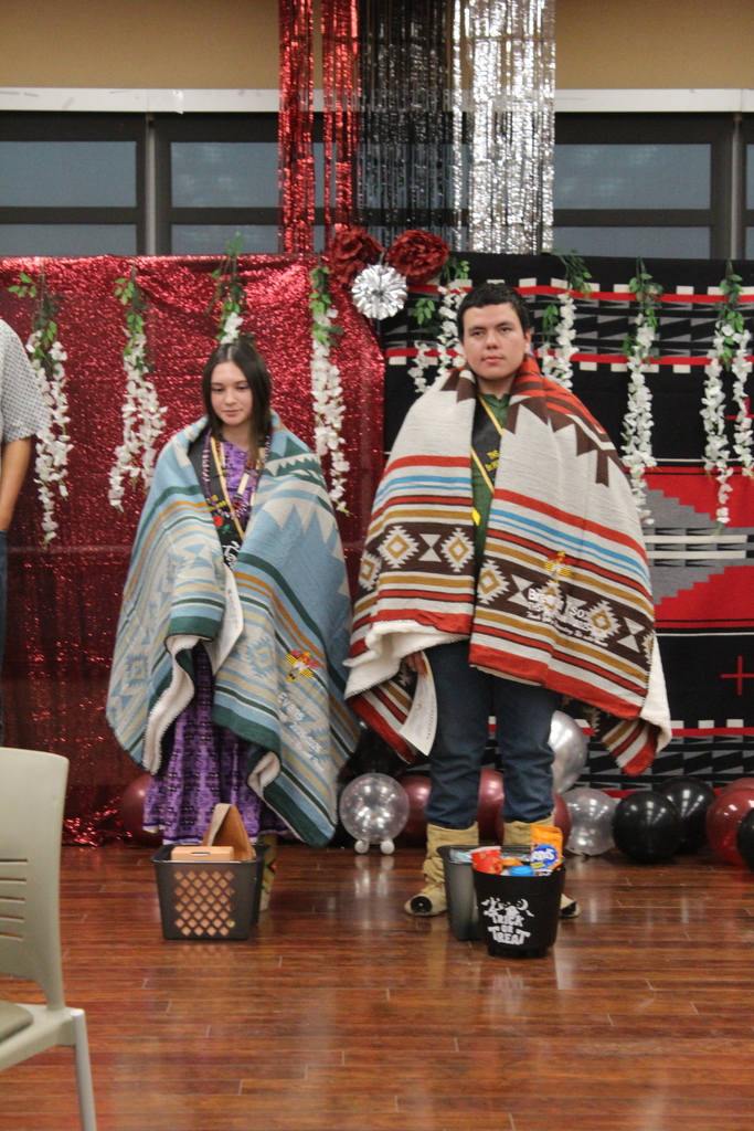 Two students stand on stage, one wearing a blue and gray blanket, the other a red, white, and black blanket.