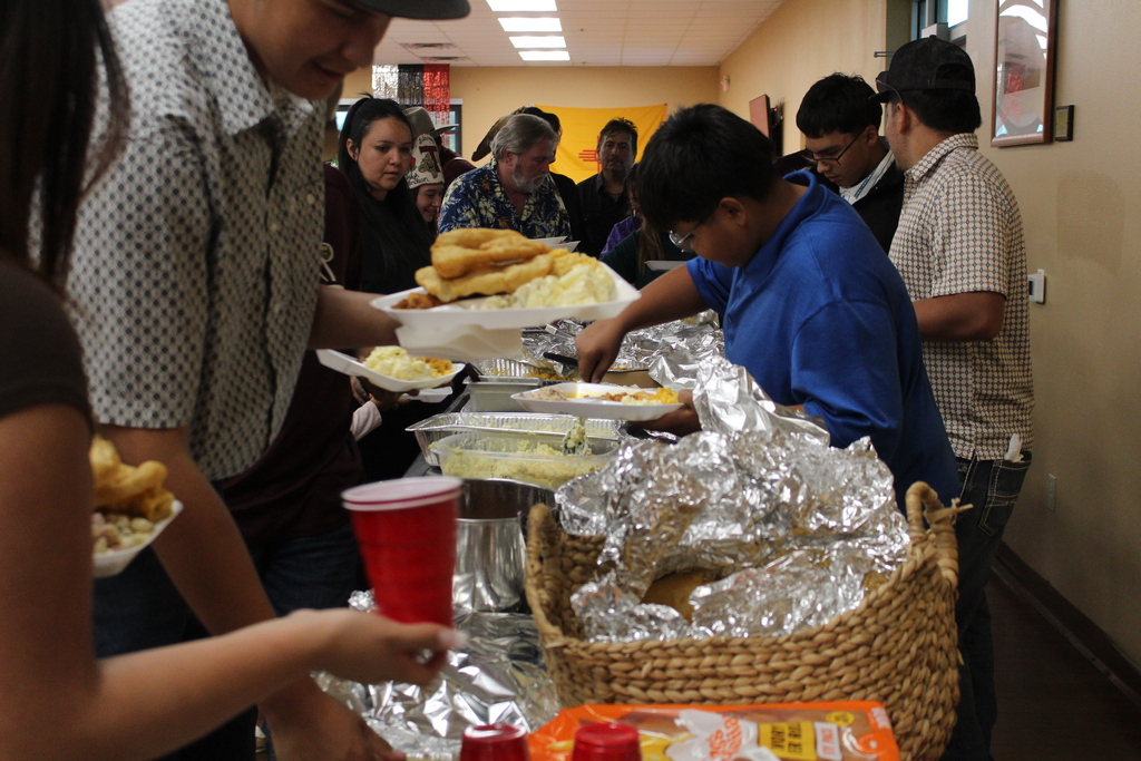 People serving and eating food at a buffet table, with covered dishes, foil wraps, and a basket.