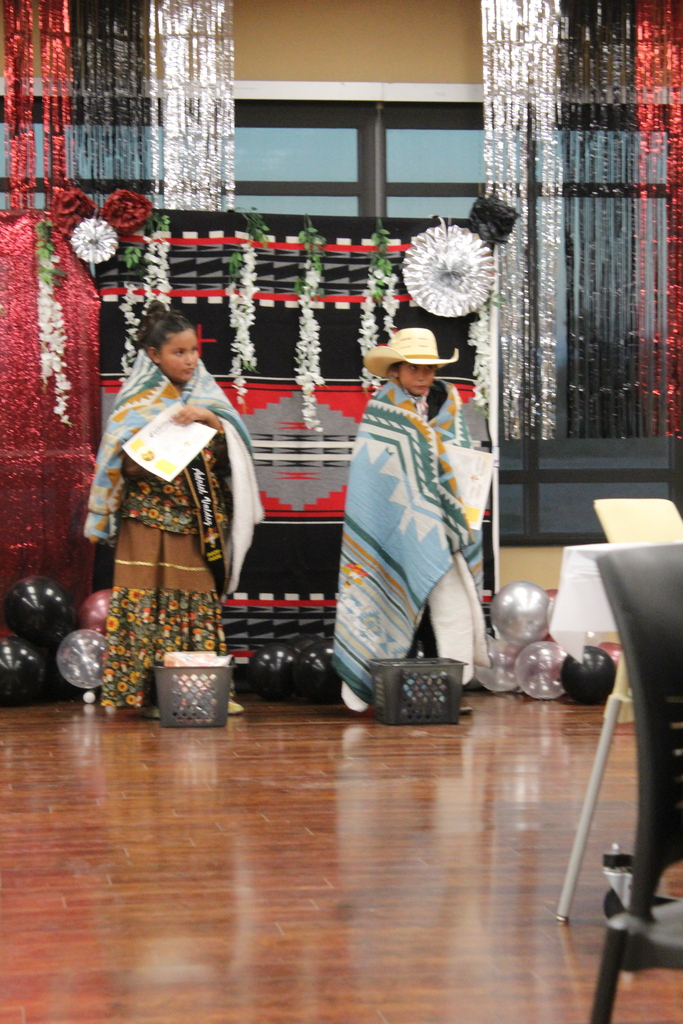 Two children in native dress stand on a stage, holding papers. Behind them is a decorative backdrop with balloons.