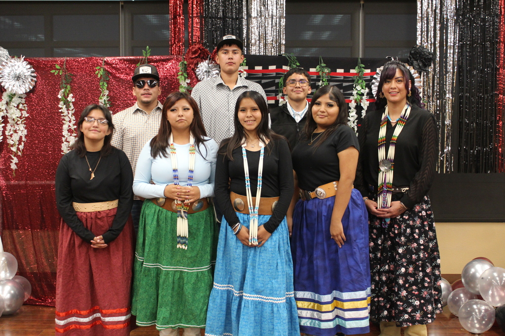 A group of students wearing traditional clothing with decorative belts and necklaces stand with their arms crossed, smiling.