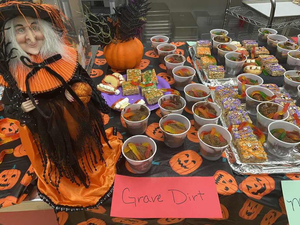 a table with an orange and black witch, black and orange tablecloth with pumpkins on it, and a tray with various Halloween lunch items