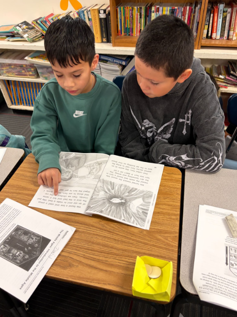 Two students sitting at a desk. One using his finger to guide him along as he read a book while the other watches him., helping as needed.