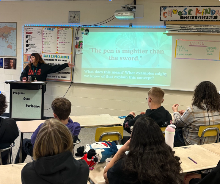 Teacher standing at podium lecturing while students watch from their desks.  