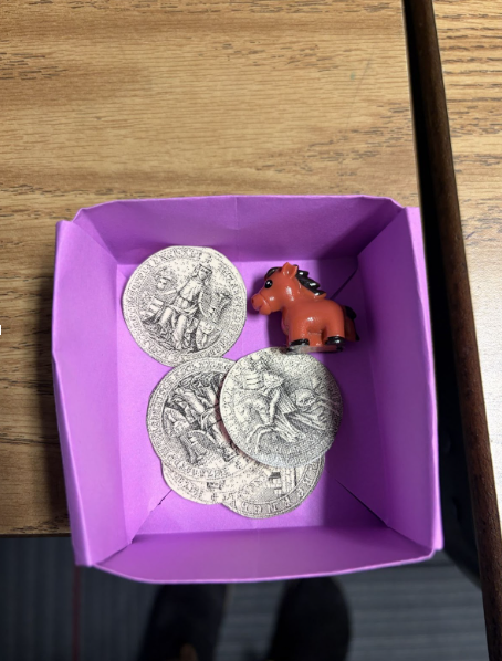 A container made out a paper sitting on a desk holding a tiny horse figuring and paper "coins" that the students are gathering to earn their Knighthood
