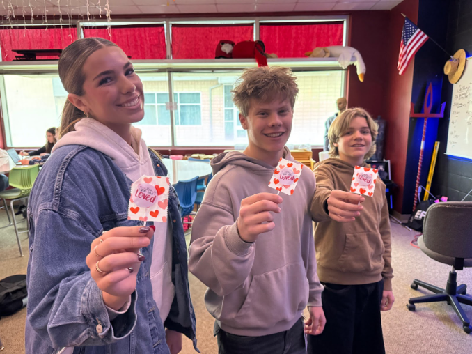 Three students smiling while holding out suckers with "You are Loved" written on the wrappers