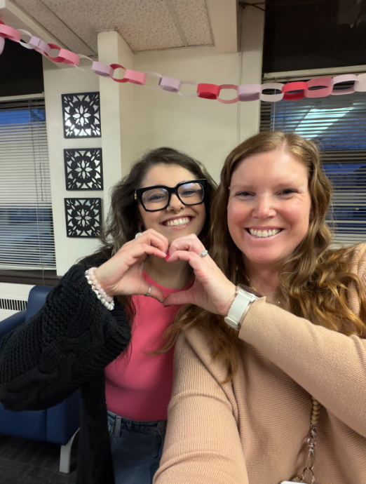 Two staff members each using one hand to gesture a heart with valentine's themed paper chain hanging behind them